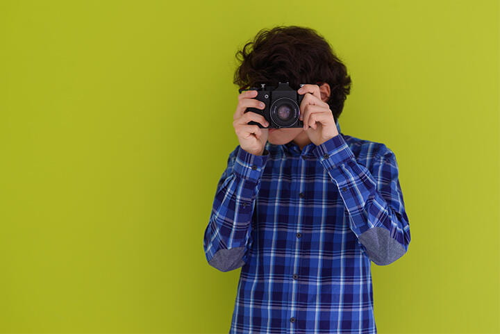 Boy standing in front on a bright green background, with a camera up to his eye.