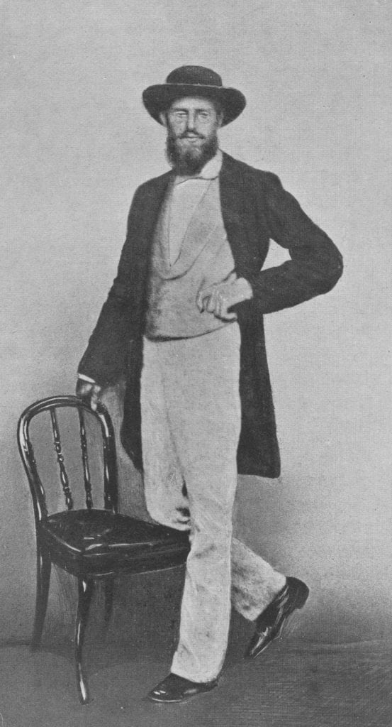 Alfred Russel Wallace wearing a hat and overcoat, standing with one knee and hand on a chair.